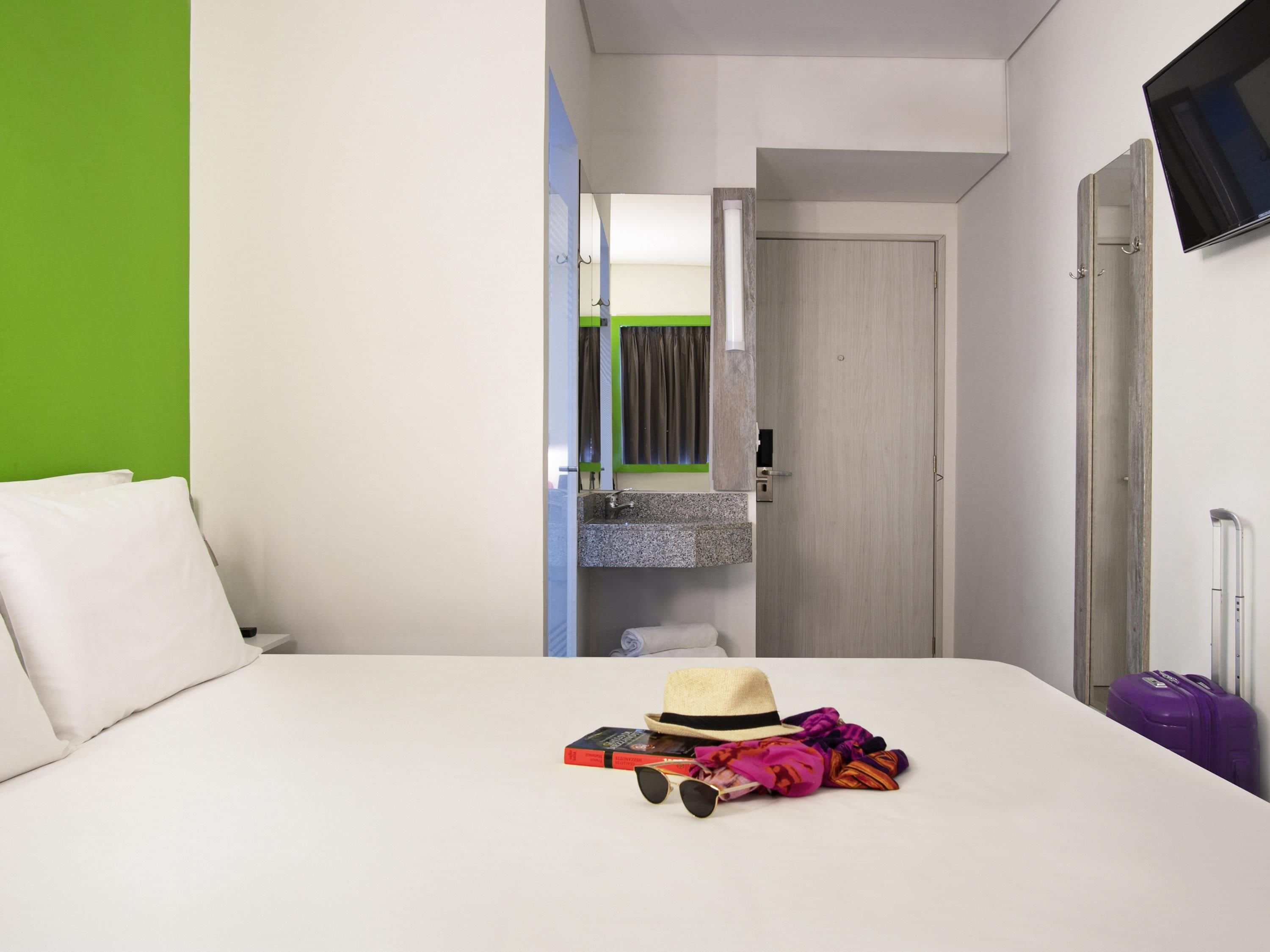 Habitación de hotel con cama, un baño pequeño con espejo, un televisor montado en la pared y una puerta que da a otra habitación.