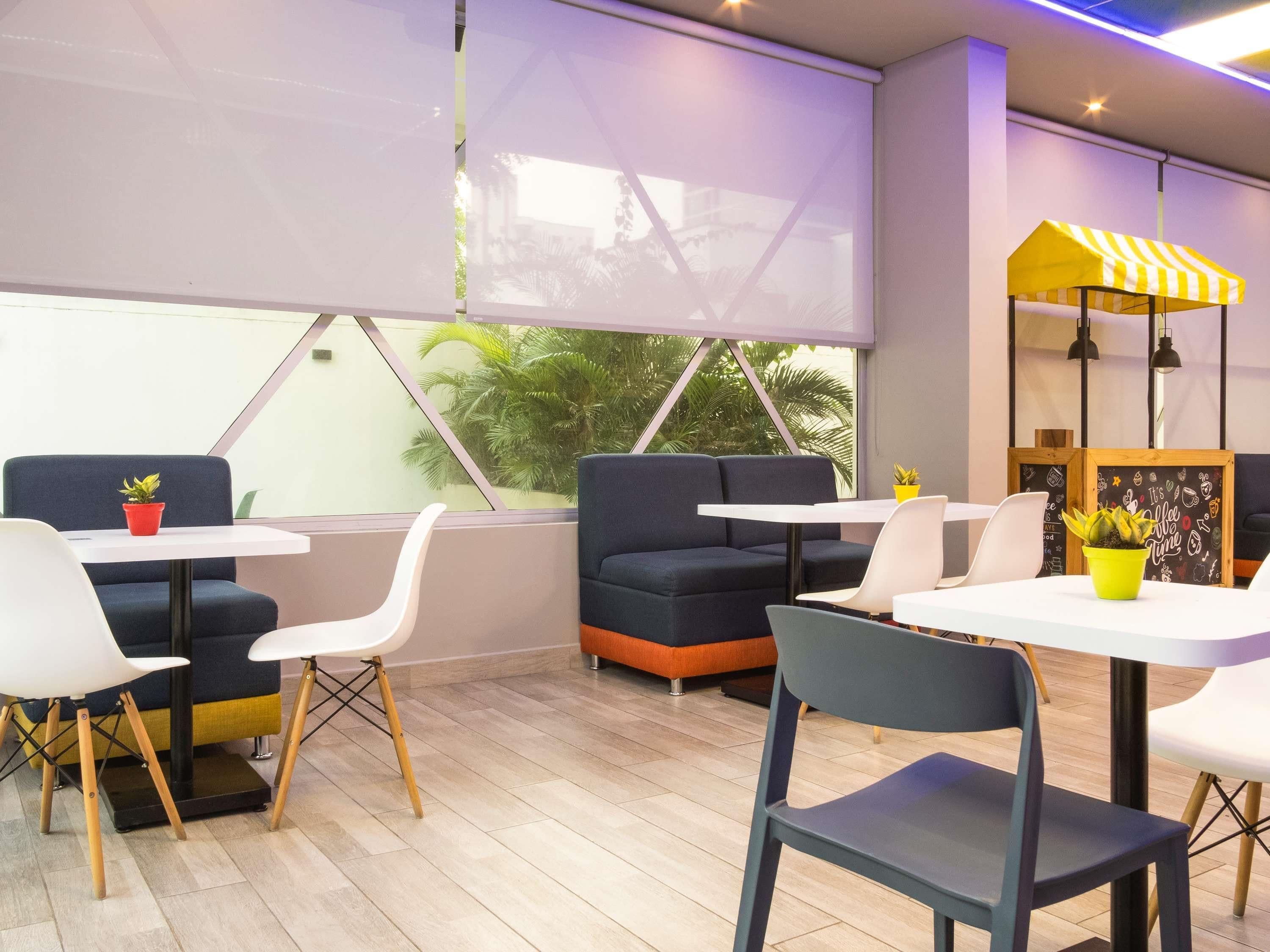 Un café moderno con disposiciones para sentarse, un toldo amarillo y blanco a rayas, y vistas a palmeras a través de grandes ventanales.
