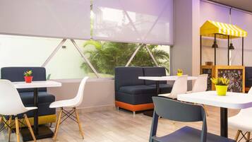 Un café moderno con disposiciones para sentarse, un toldo amarillo y blanco a rayas, y vistas a palmeras a través de grandes ventanales.