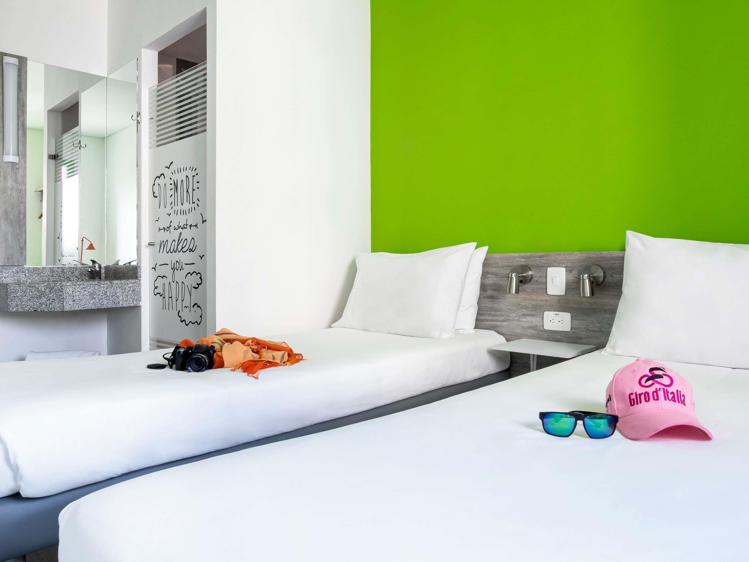 Habitación de hotel con dos camas, una pared de acento verde, un espejo y una lámpara fijada a la pared.