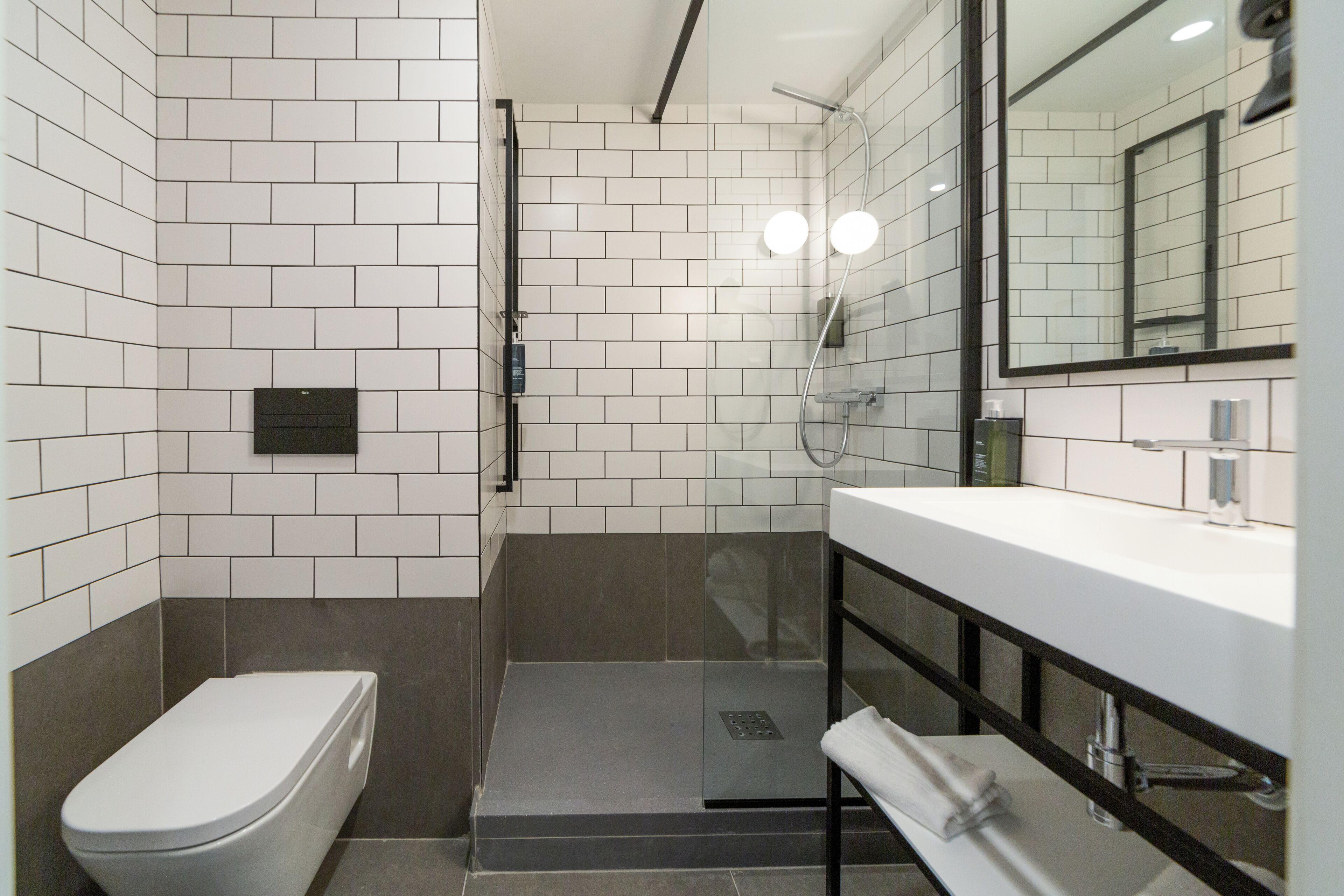 Une salle de bain moderne avec des murs recouverts de carreaux blancs, des toilettes, une douche et un lavabo avec un miroir.