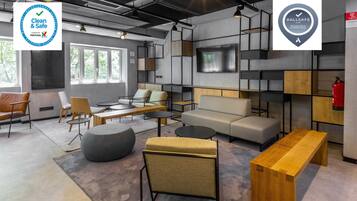 A modern hotel lobby with a clean and safe environment, featuring a sofa, chairs, a wooden bench, and a TV mounted on the wall.