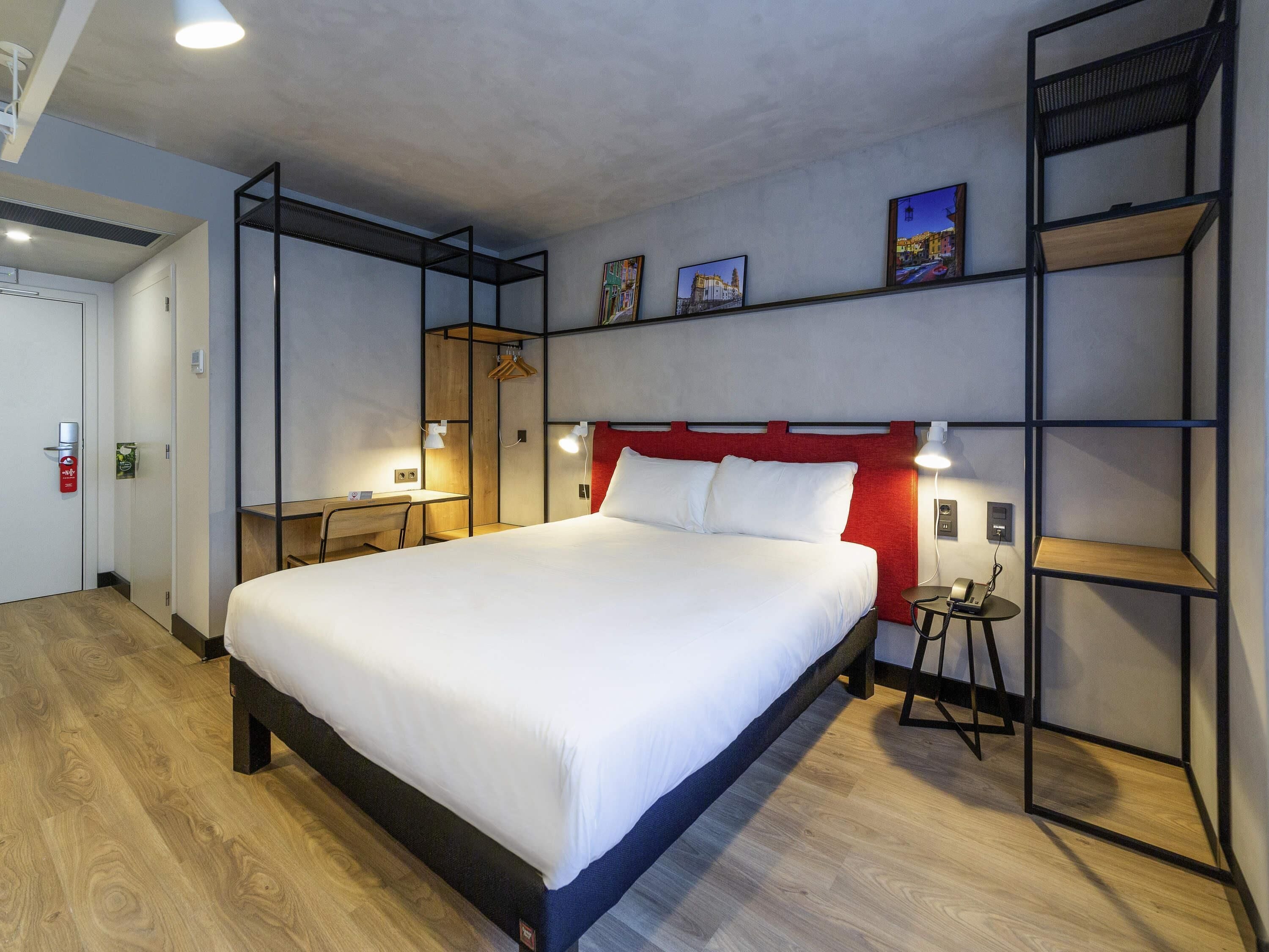 Una habitación de hotel moderna con una cama grande, un escritorio y un armario. Hay estantes en la pared y una pequeña mesa con una lámpara.