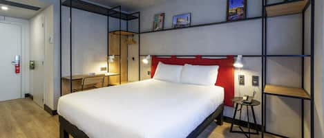 Una habitación de hotel moderna con una cama grande, un escritorio y un armario. Hay estantes en la pared y una pequeña mesa con una lámpara.