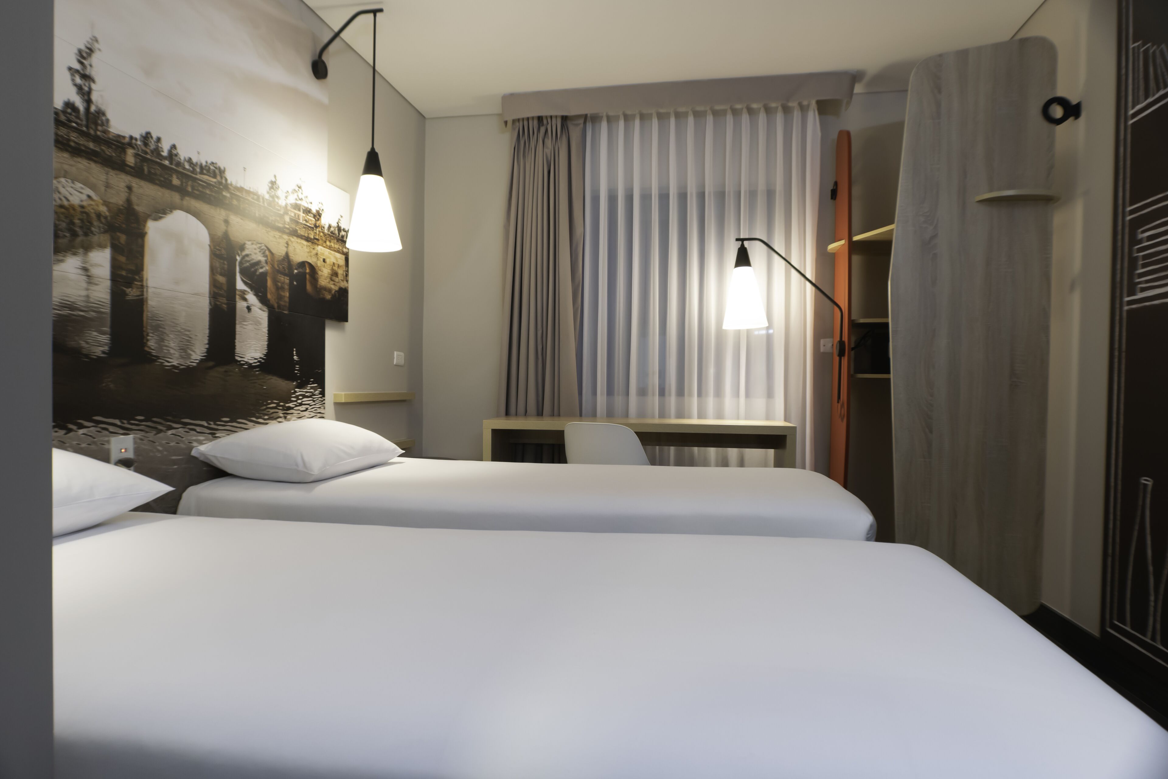 A hotel room with two beds, a desk, a chair, a painting on the wall, and a window with curtains.