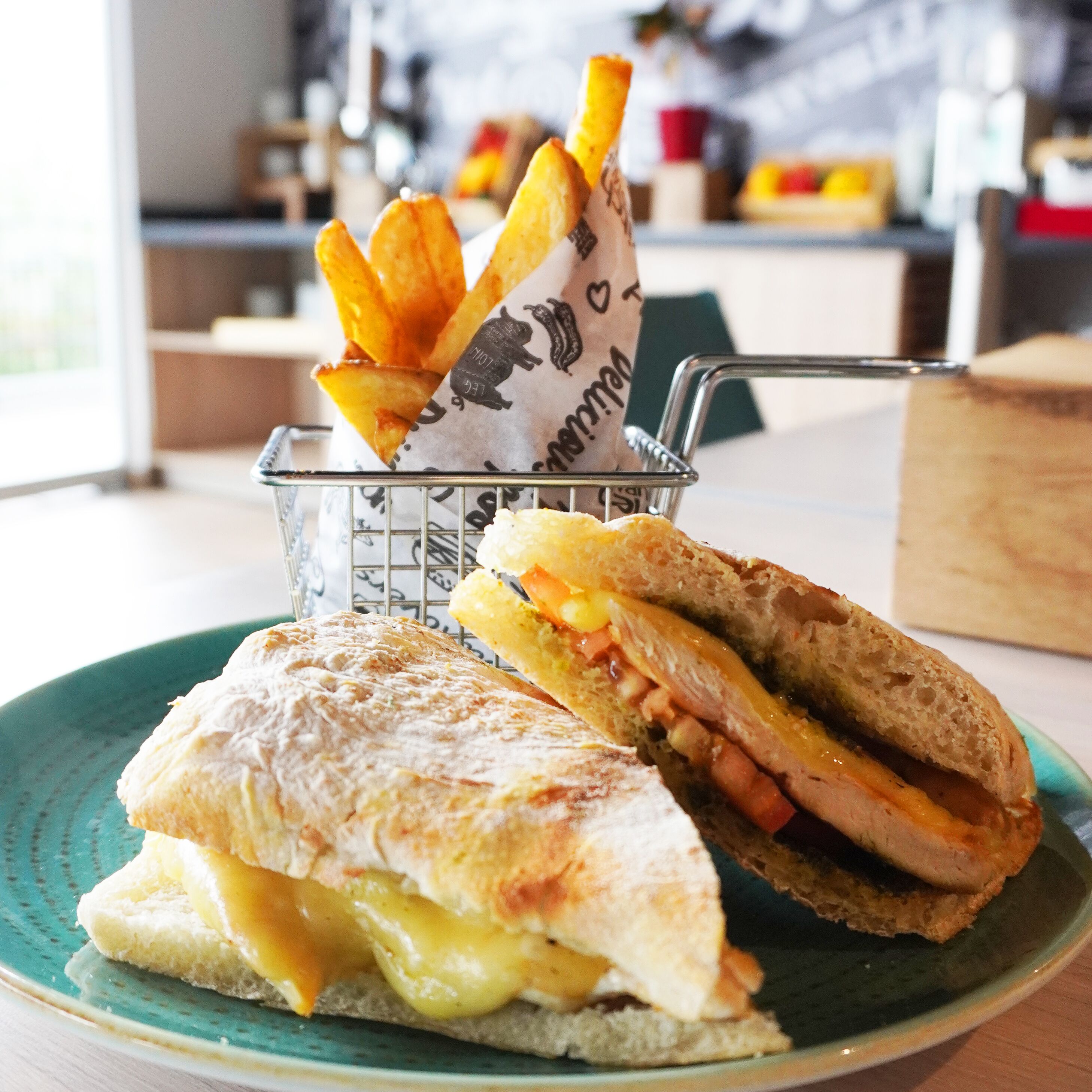 A sandwich with cheese and ham, a plate of fries, and a basket with a paper bag.