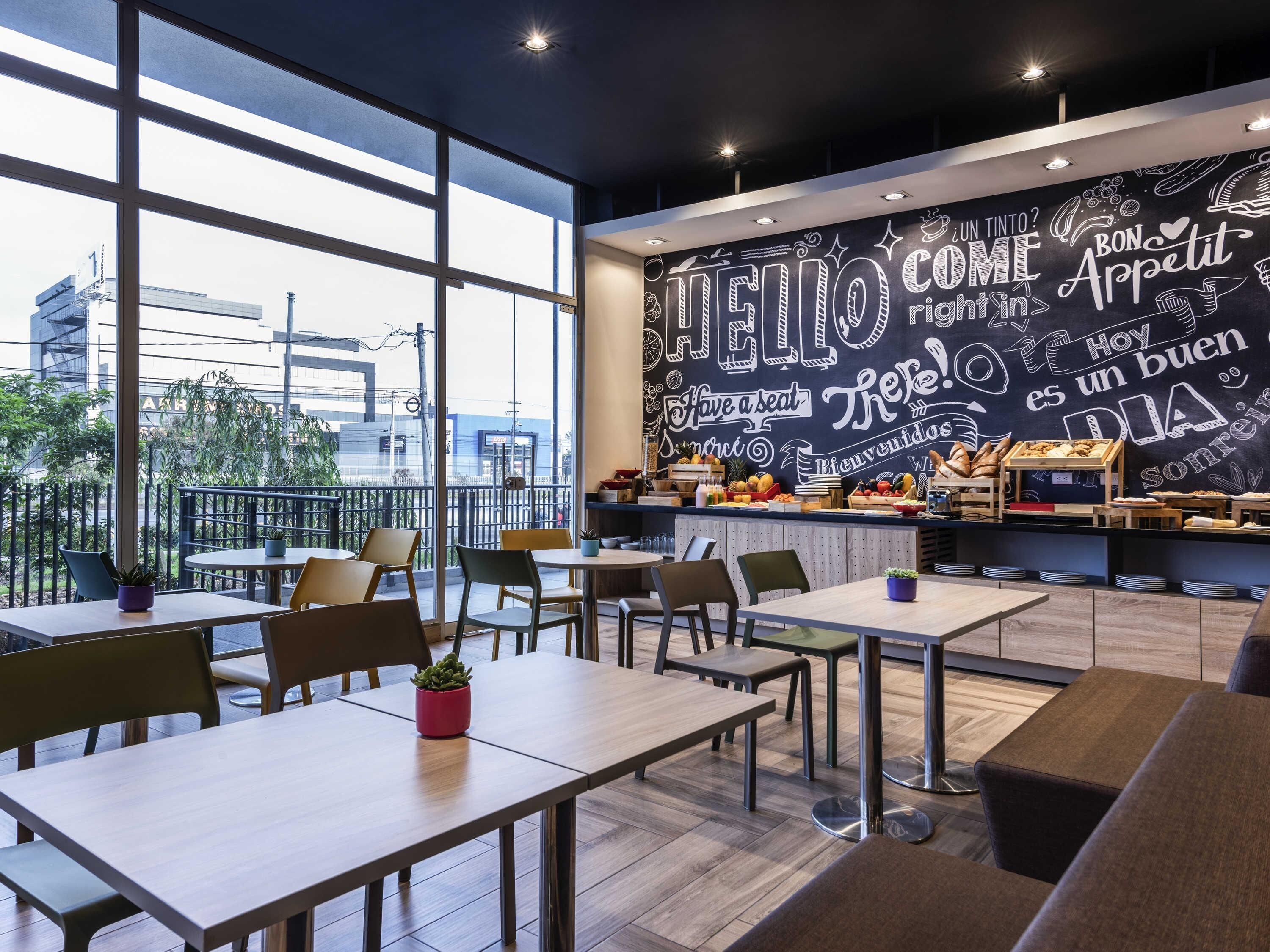A cafe with a large window, tables, chairs, and a chalkboard wall with various words and phrases.