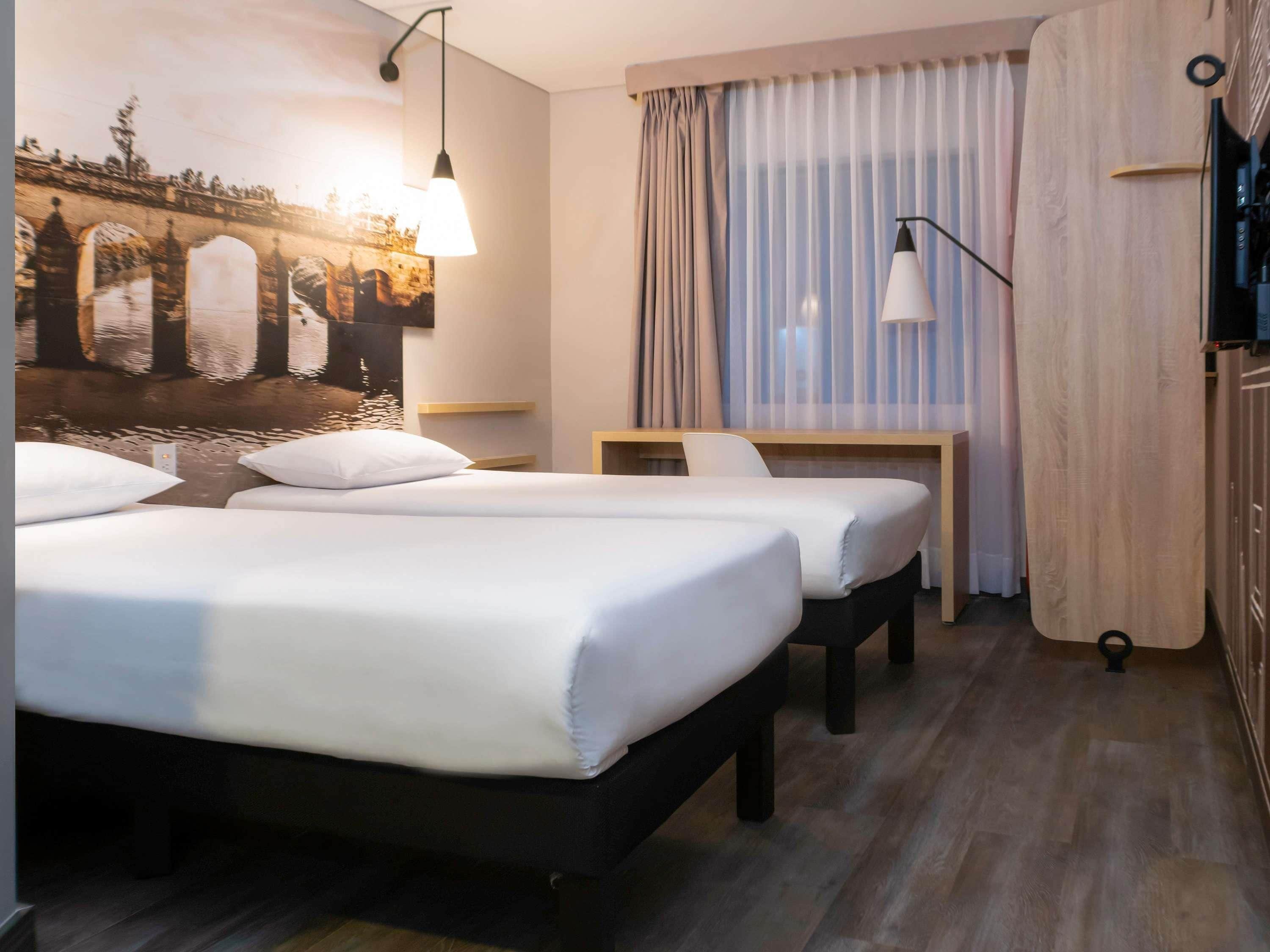 Habitación de hotel con dos camas, un escritorio, una silla, televisión y un ventanal con cortinas.