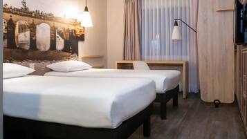 Habitación de hotel con dos camas, un escritorio, una silla, televisión y un ventanal con cortinas.