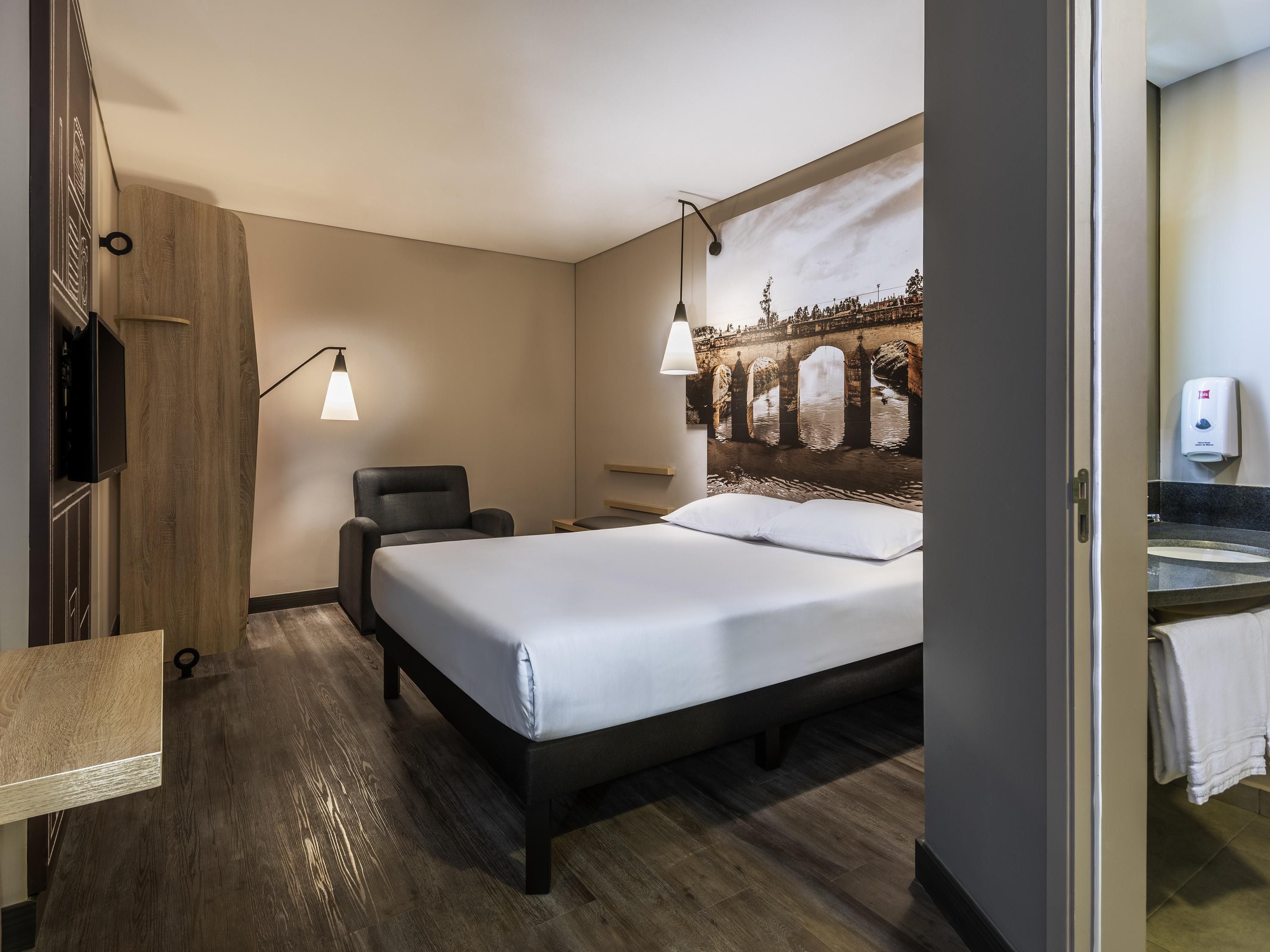 A hotel room with a bed, a chair, a desk, a wall-mounted TV, and a painting of a bridge.