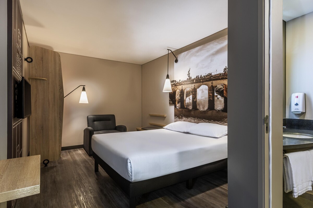 A hotel room with a bed, a chair, a desk, a wall-mounted TV, and a painting of a bridge.