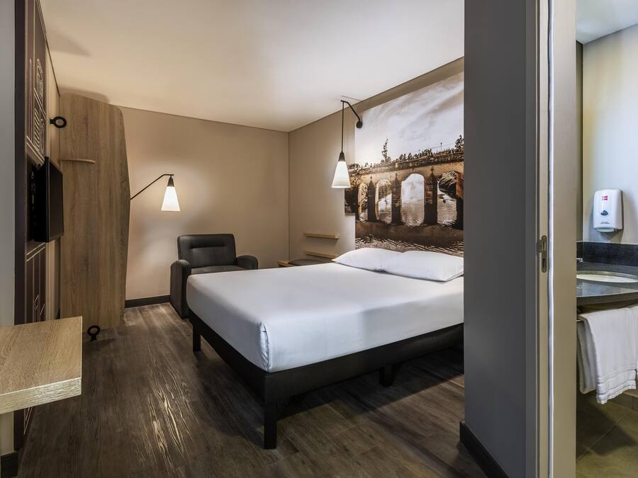 A hotel room with a bed, a chair, a desk, a wall-mounted TV, and a painting of a bridge.