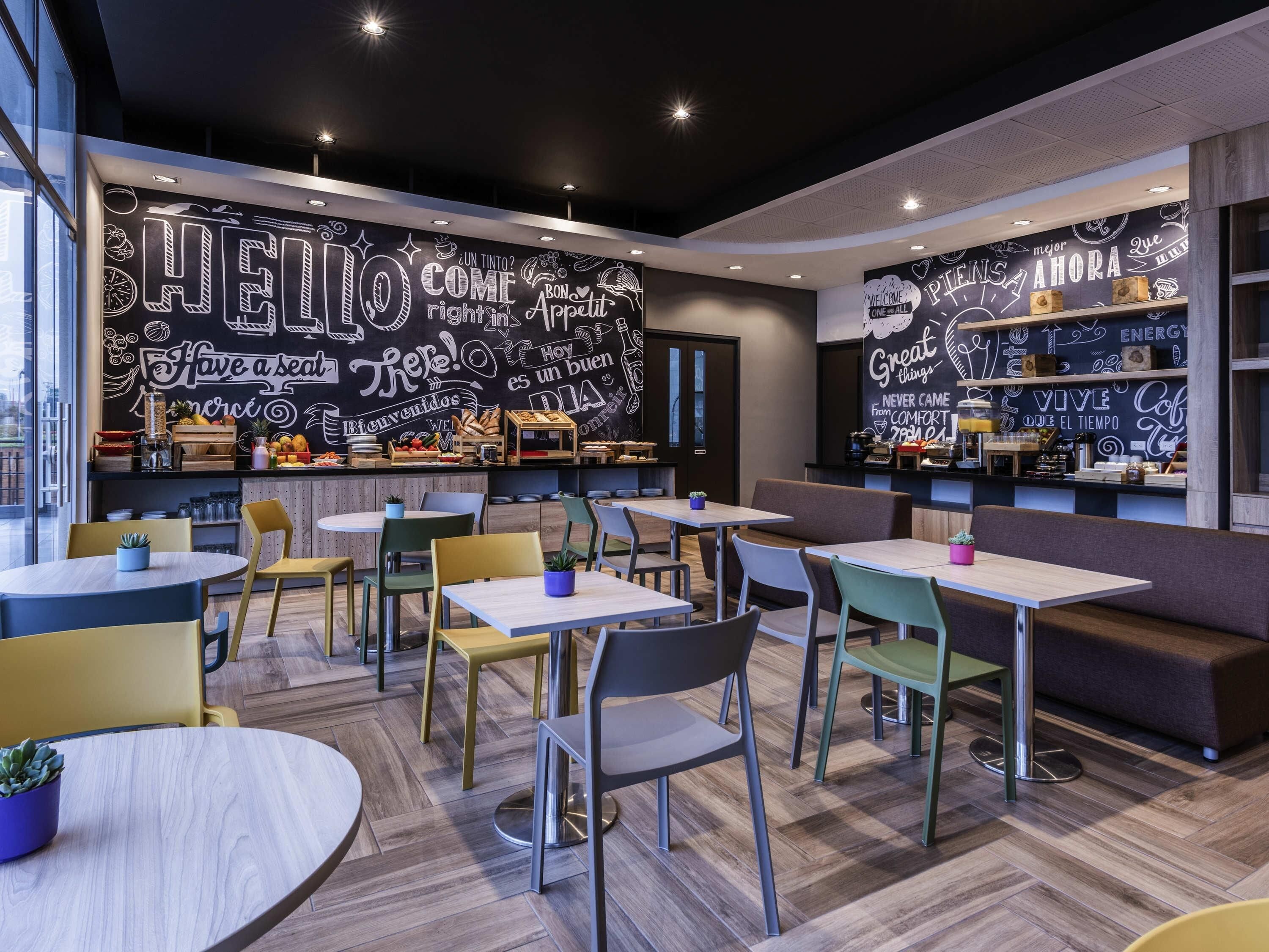 Interior moderno de una cafetería con una pared de pizarra que muestra diversas frases y una zona de bar con estantes llenos de aperitivos y bebidas.
