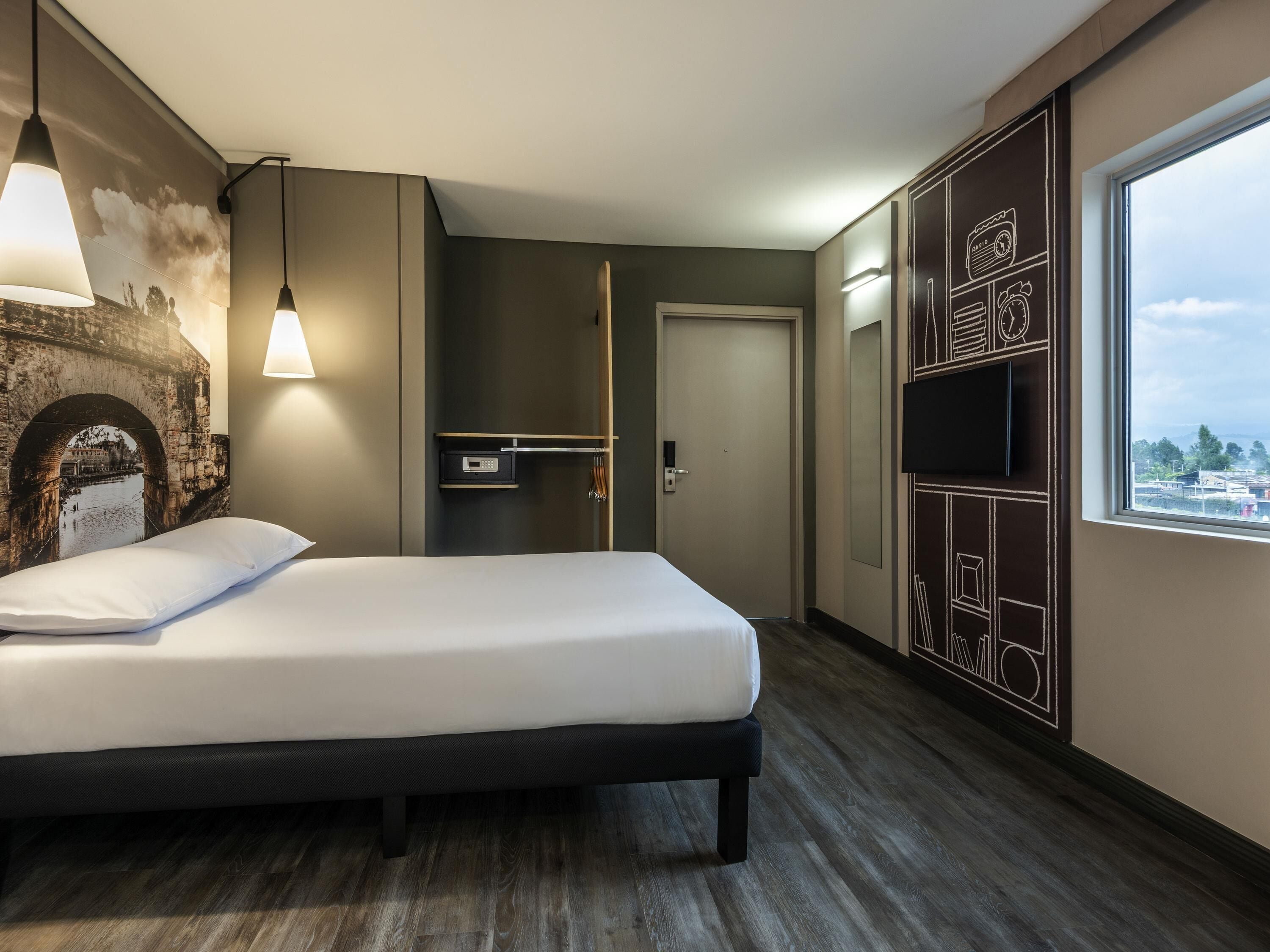 A modern hotel room with a bed, a wall-mounted TV, a wardrobe, and a large window.