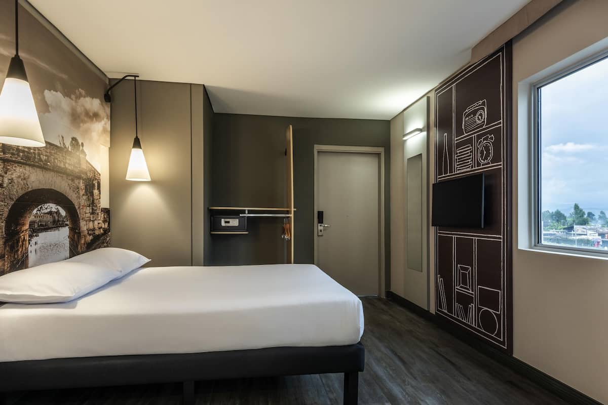 A modern hotel room with a bed, a wall-mounted TV, a wardrobe, and a large window.