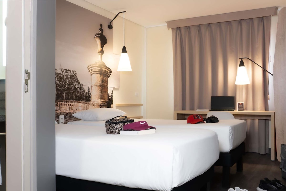 Habitació d'hotel amb dos llits, un escriptori amb un ordinador i un mural gran a la paret.