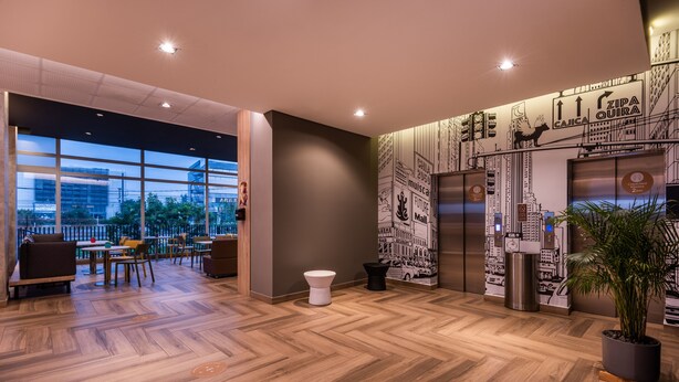 A modern lobby with wooden flooring, a seating area with tables and chairs, a large window, and a wall with cityscape illustrations. - ibis Chia (Chia)