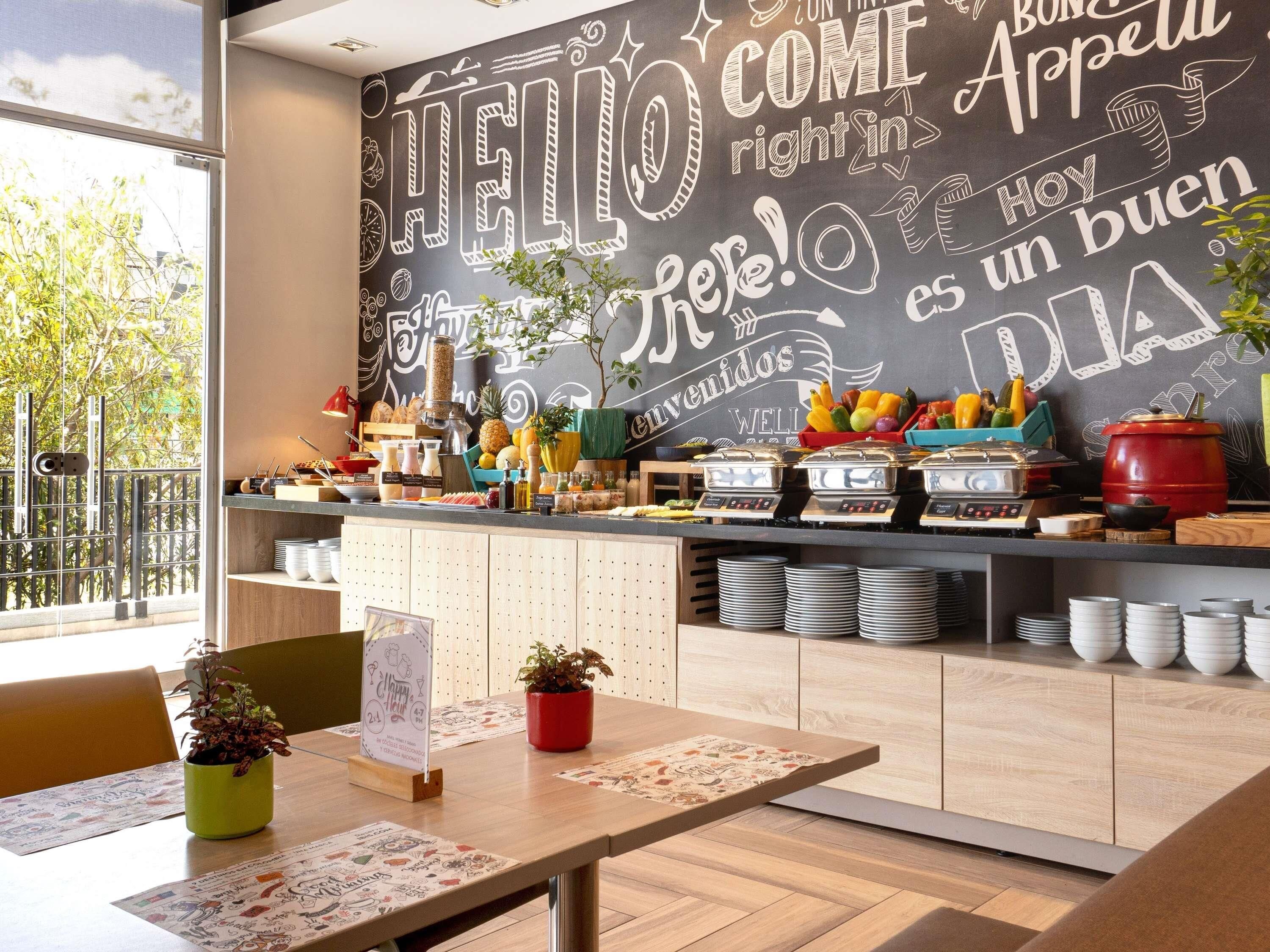 Una cafetería con una pared de pizarra que muestra varios saludos y un mostrador de buffet con alimentos.