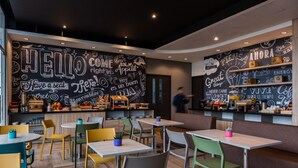 A modern cafe with a chalkboard wall featuring various phrases and doodles, wooden tables and chairs, and a counter with food items. - ibis Chia (Chia)