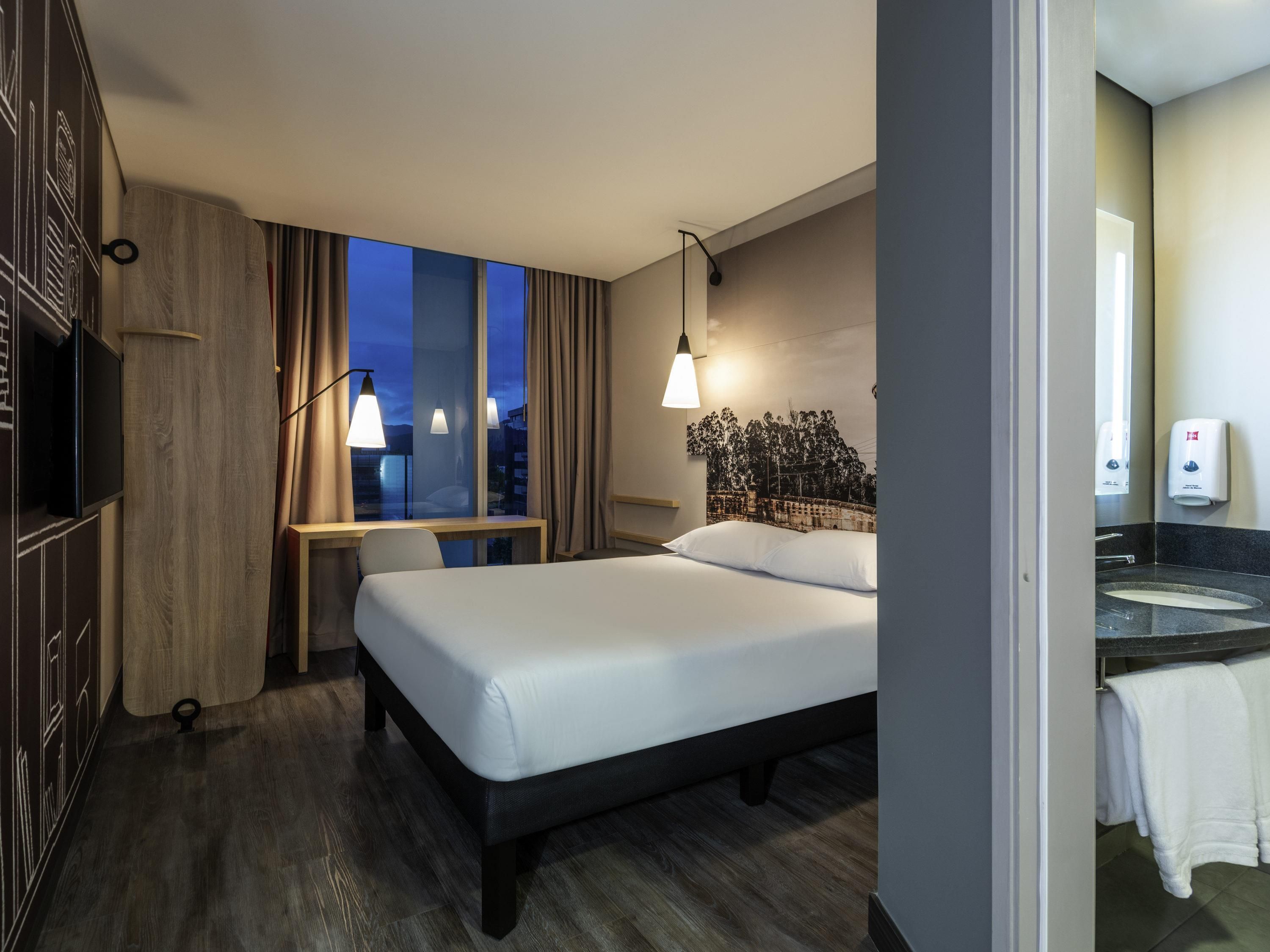 Quarto de hotel com cama, escrivaninha, televisão, janela com cortinas, luminária na parede e banheiro com pia e porta-toalhas.