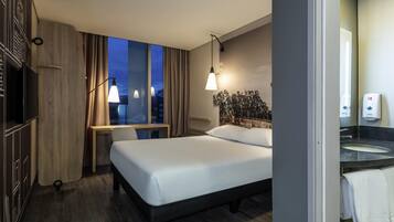 Quarto de hotel com cama, escrivaninha, televisão, janela com cortinas, luminária na parede e banheiro com pia e porta-toalhas.