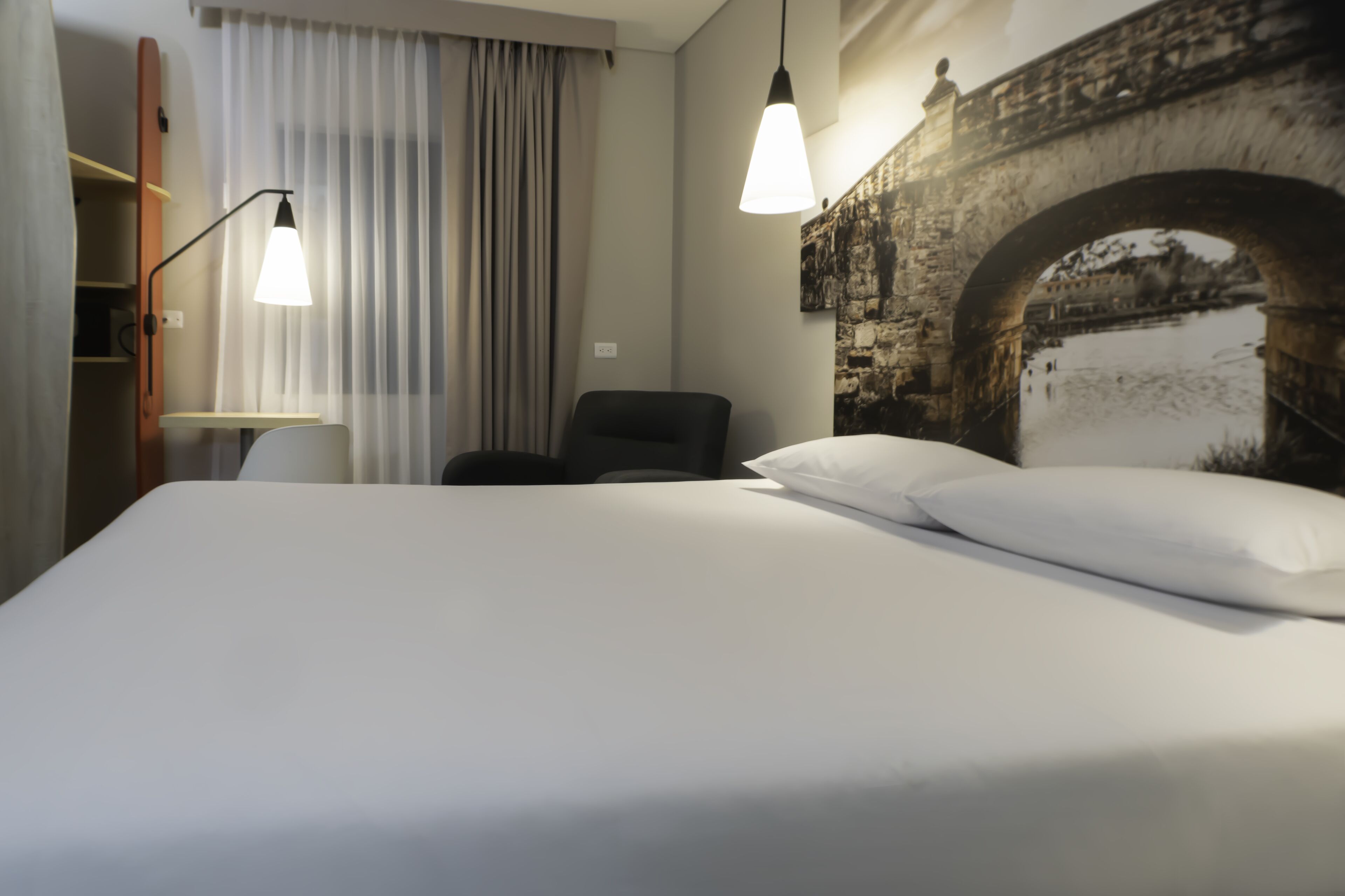 Habitación de hotel con cama, una silla, un escritorio y un amplio ventanal con cortinas.