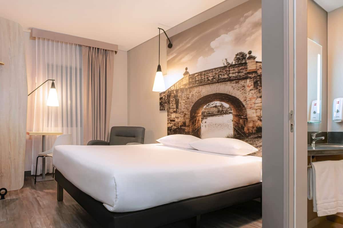 Una habitación de hotel con cama, una silla, un escritorio, una lámpara, un baño con espejo y un mural de un puente.