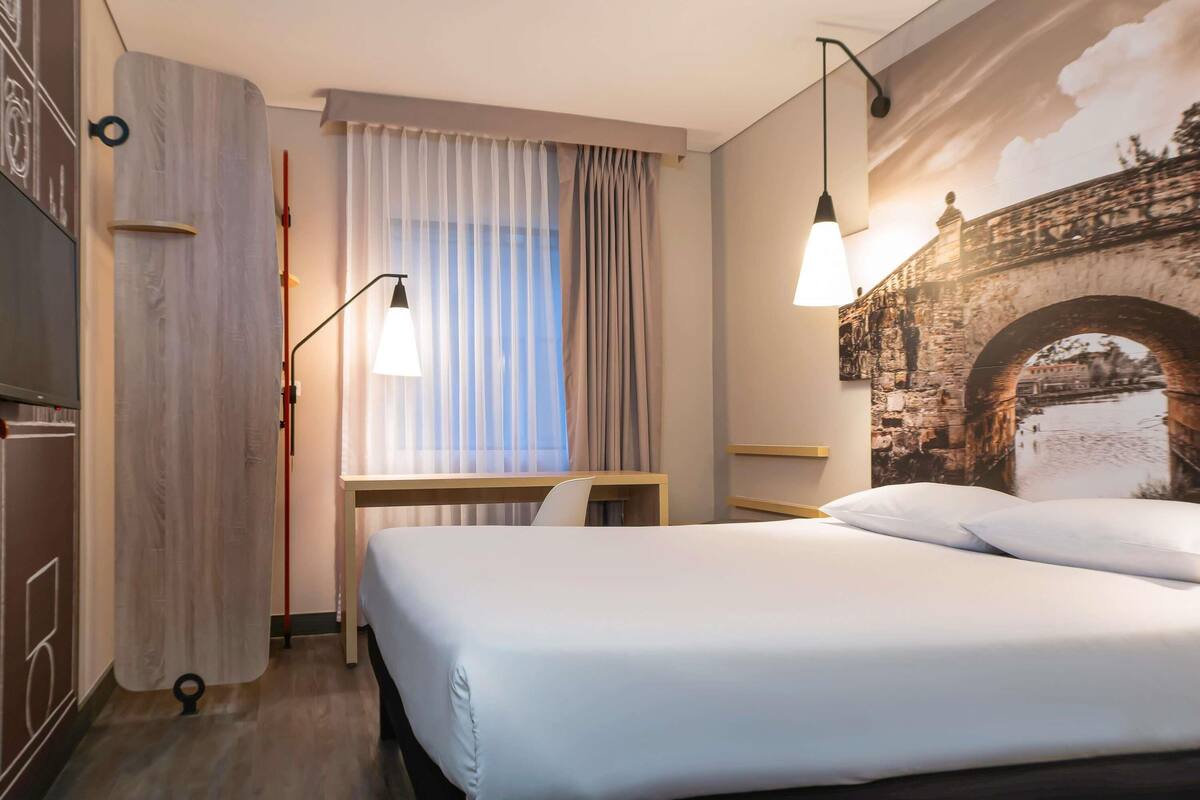 A hotel room with a bed, a desk, a chair, a lamp, and a mural of a bridge.