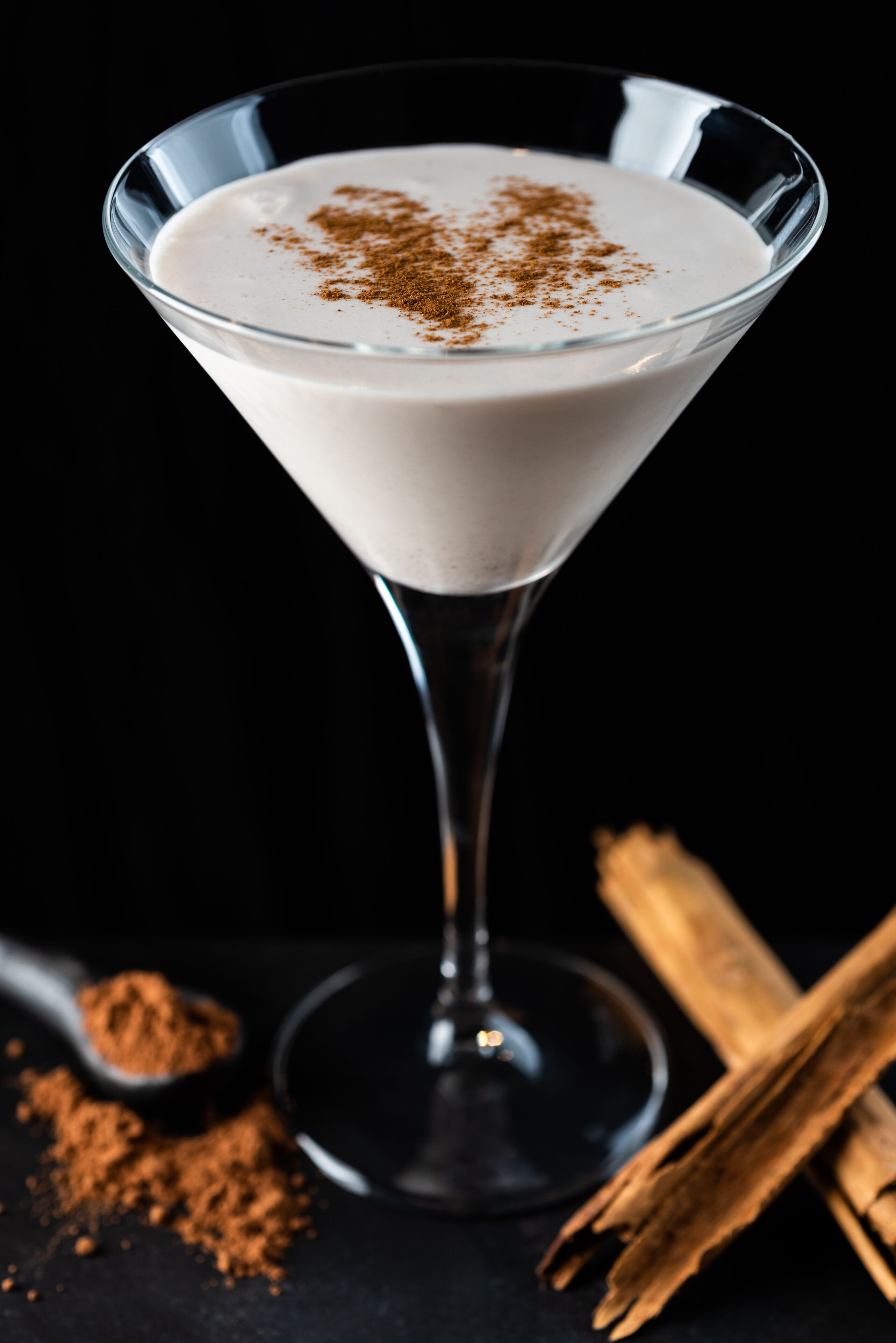 Ein Martini-Glas mit einem cremigen Getränk, bestäubt mit braunem Pulver, vermutlich Zimt, vor dunklem Hintergrund.