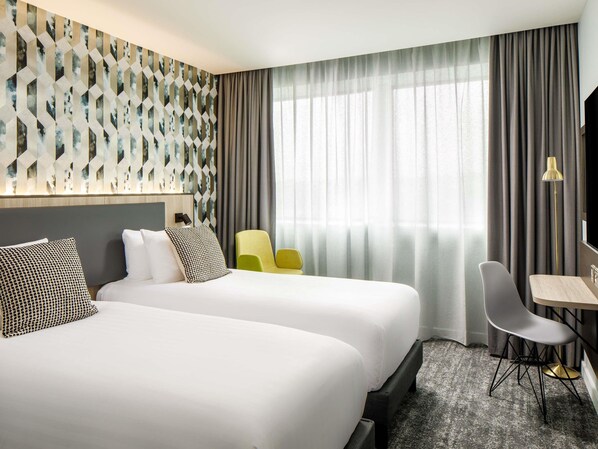 Habitación de hotel con dos camas, un escritorio, una silla, una lámpara y un ventanal grande con cortinas.