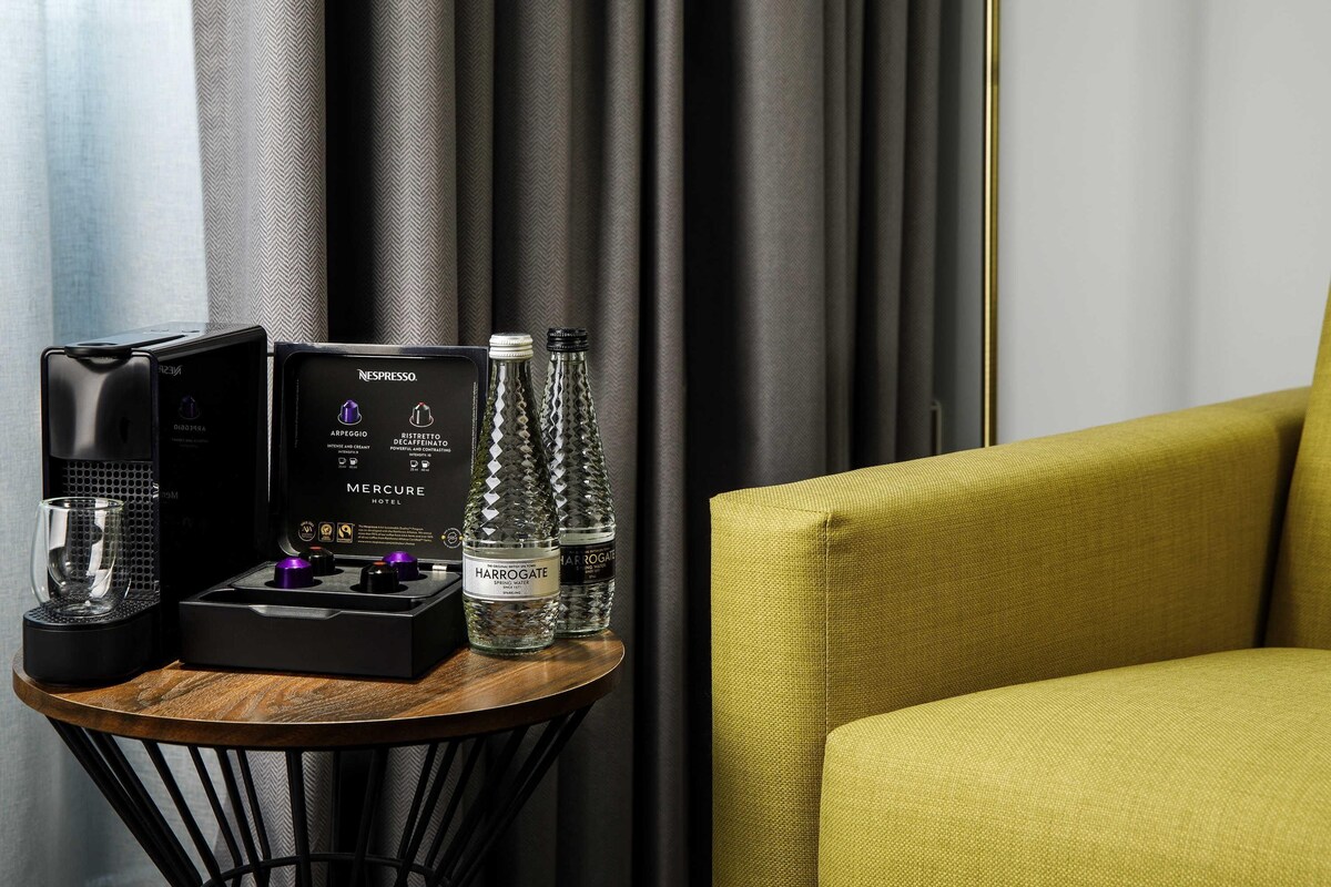 A minibar setup with a coffee machine, a water bottle, and a glass on a wooden table, next to a yellow armchair.