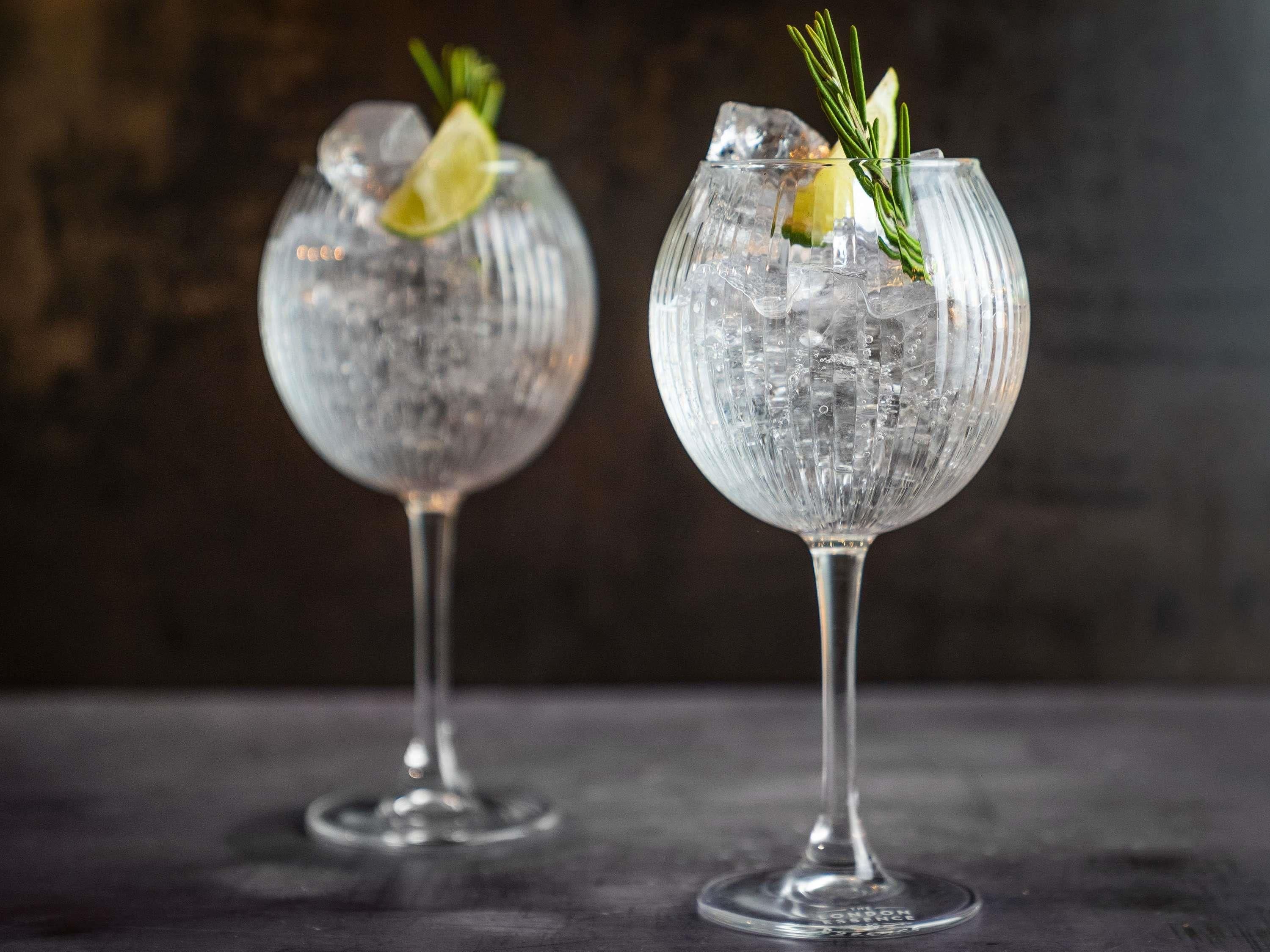 Two glasses of a clear beverage with ice cubes and garnishes, placed on a dark surface.