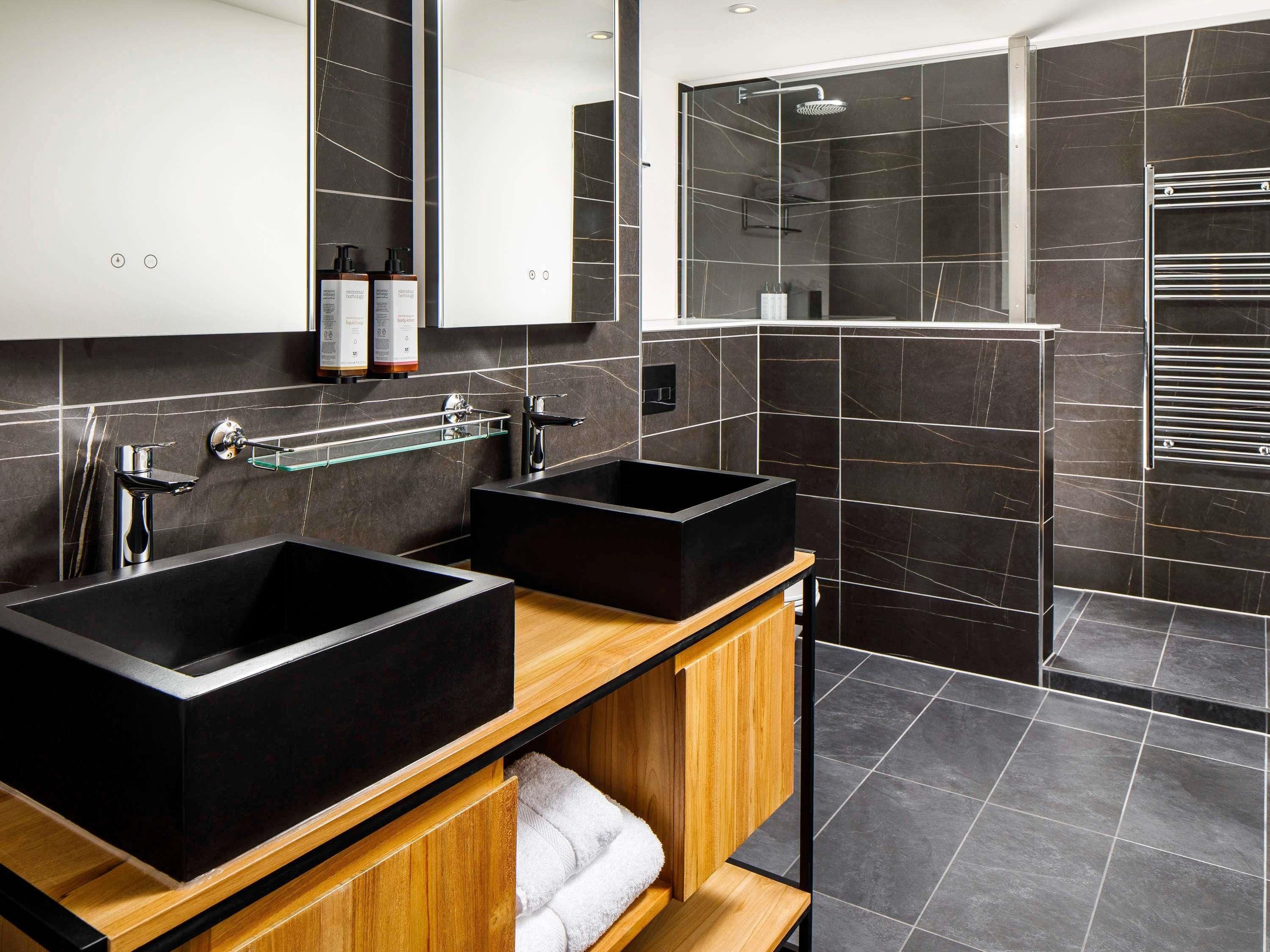 A modern bathroom with two black sinks, a wooden vanity, a glass shelf, and a shower area.