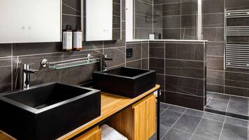 A modern bathroom with two black sinks, a wooden vanity, a glass shelf, and a shower area.