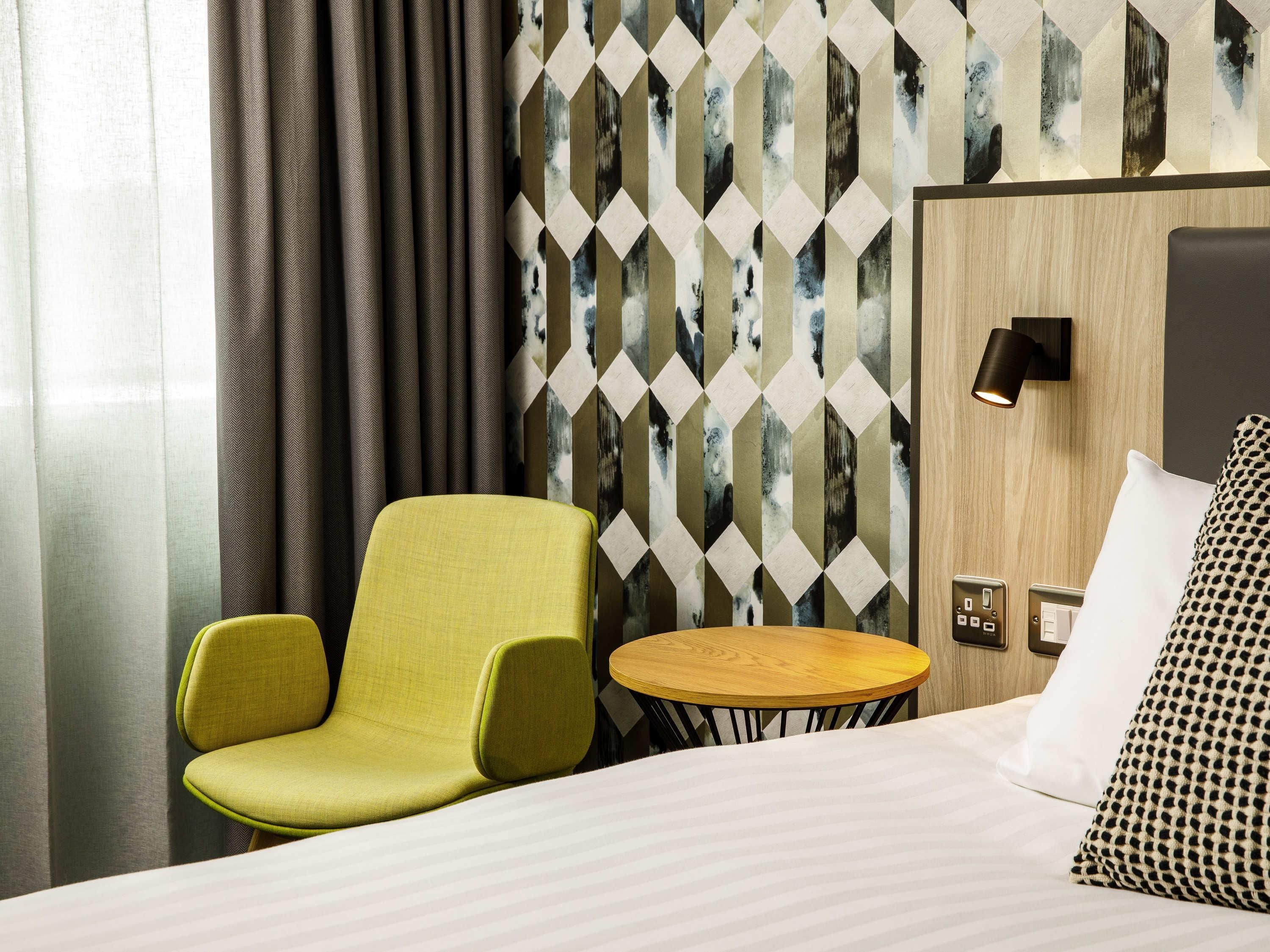 A hotel room with a bed, a green armchair, a small round table, a wall-mounted lamp, and a patterned wallpaper.
