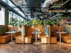 A modern restaurant interior with wooden booths, round tables, and green plants. - Mercure Newport Hotel (Newport)