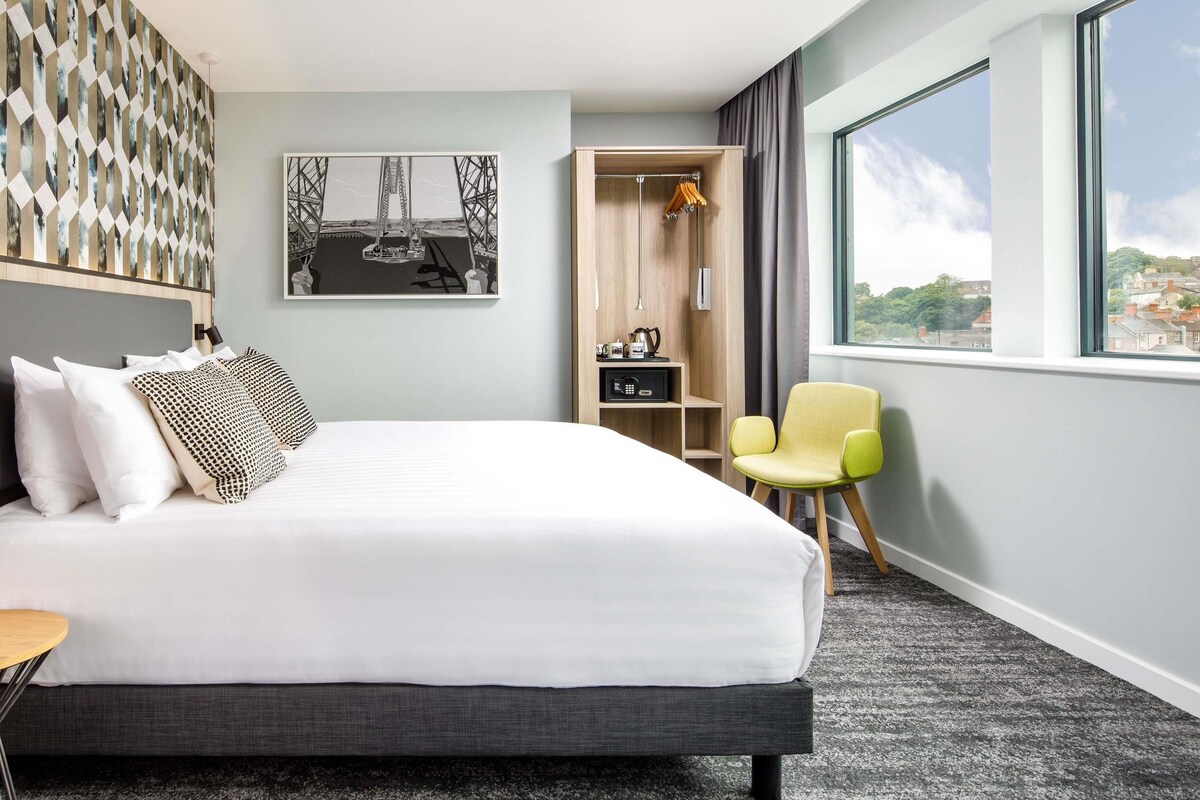 A hotel room with a bed, a chair, a wardrobe, a desk, and a large window.