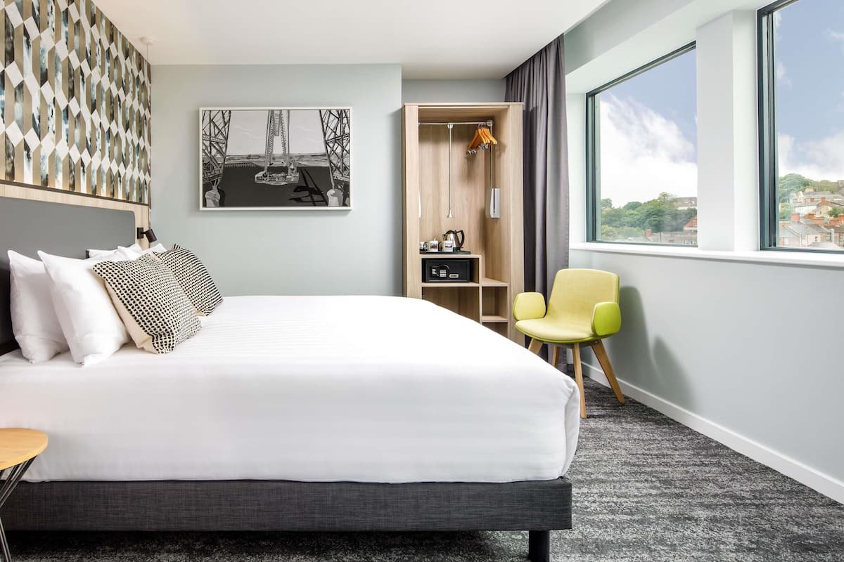 A hotel room with a bed, a chair, a wardrobe, a desk, and a large window.