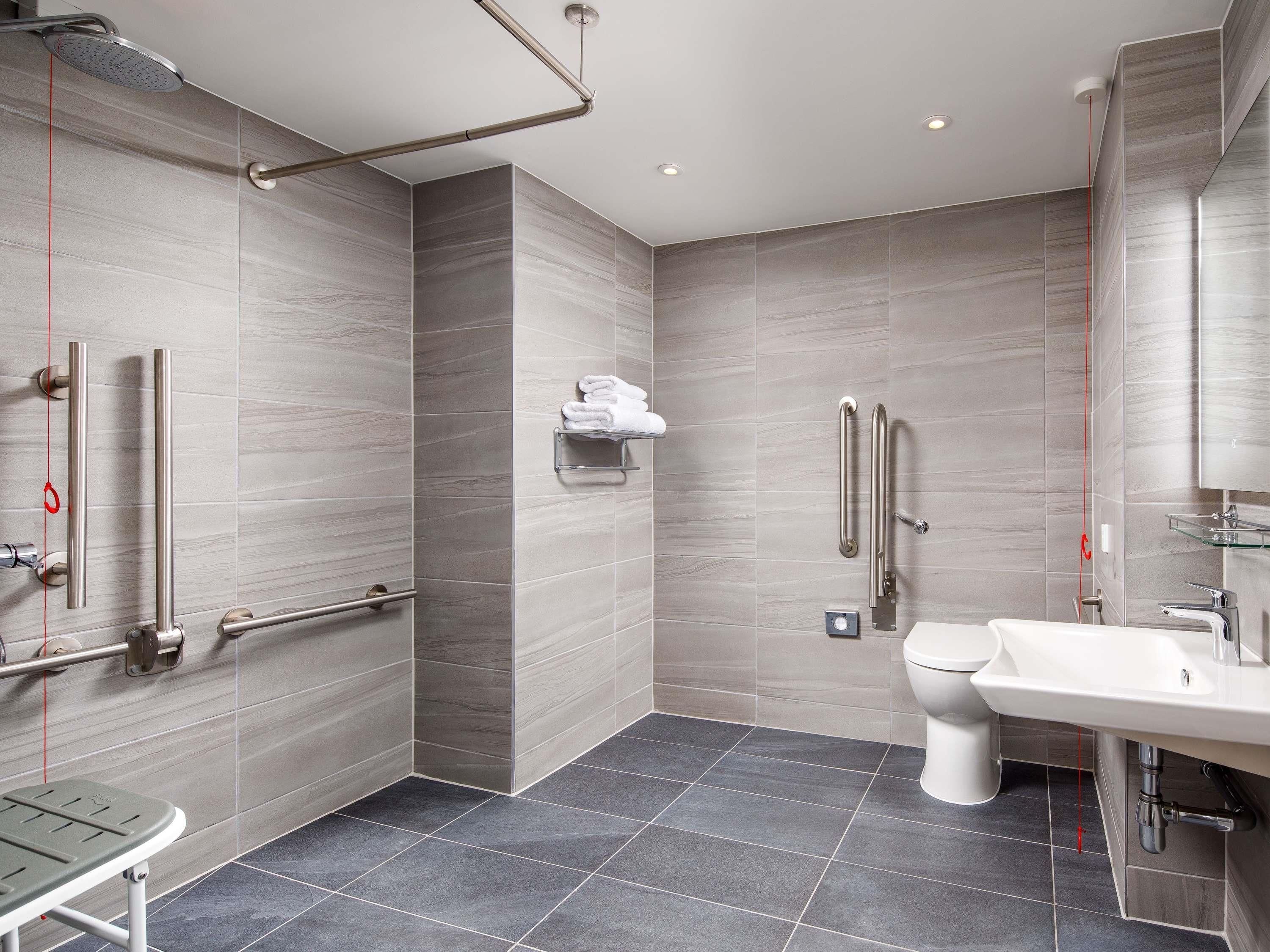 Un baño moderno con ducha, inodoro y lavamanos, equipado con barras de apoyo y un perchero para toallas.