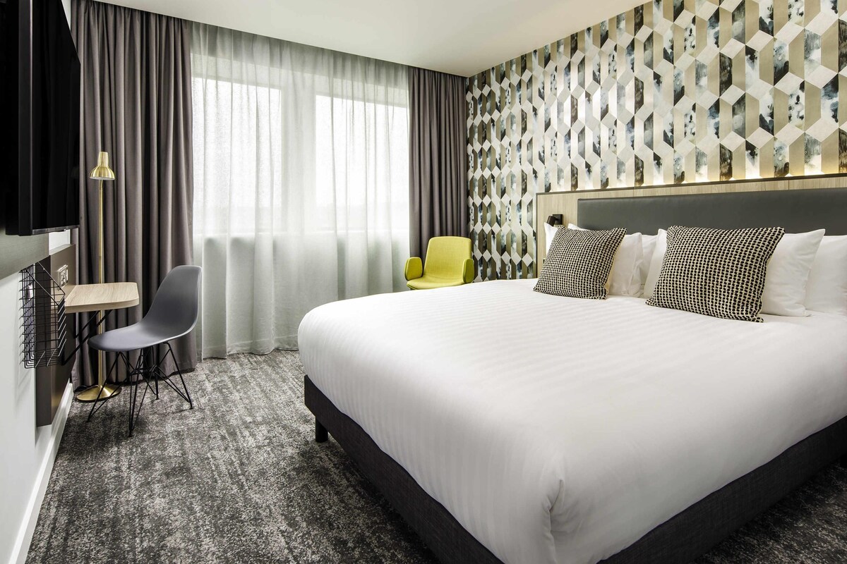 A hotel room with a large bed, a desk with a chair, a TV, and a patterned wall.