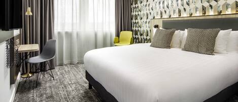 A hotel room with a large bed, a desk with a chair, a TV, and a patterned wall.