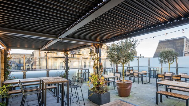 An outdoor patio area with tables and chairs, potted plants, and a view of buildings in the background. - Mercure Newport Hotel (Newport)