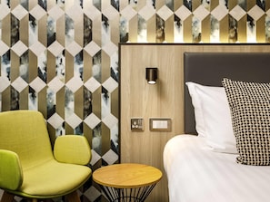 A hotel room with a geometric patterned wall, a wooden headboard, a bed with white linens, a green armchair, a small wooden table, and a wall-mounted light fixture. - Mercure Newport Hotel (Newport)