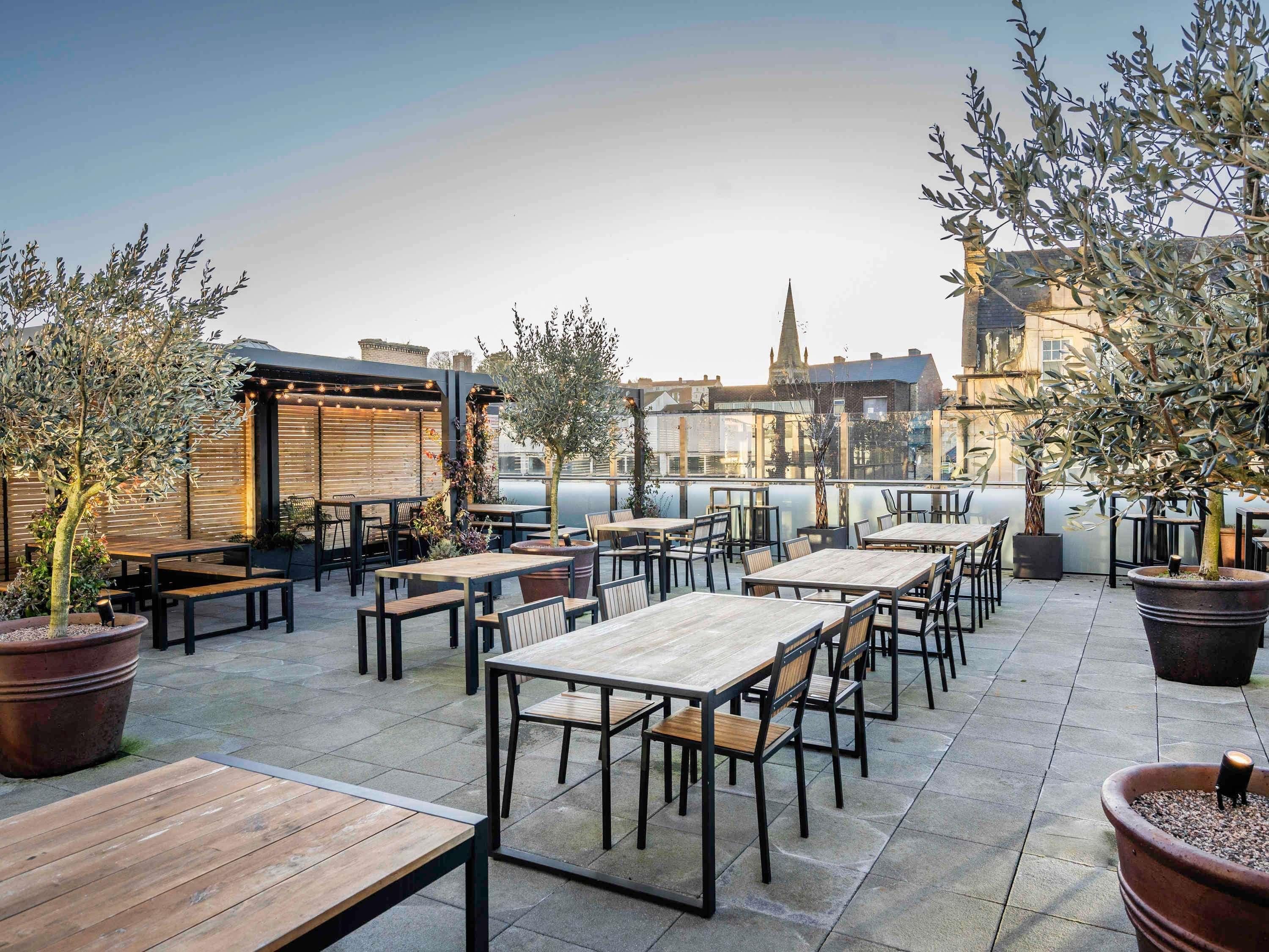 Une terrasse extérieure avec des tables et des chaises, des plantes en pot et une vue sur les bâtiments en arrière-plan.