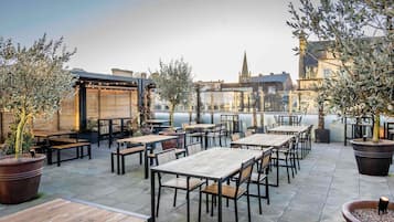 An outdoor patio with tables and chairs, potted plants, and a view of buildings in the background.