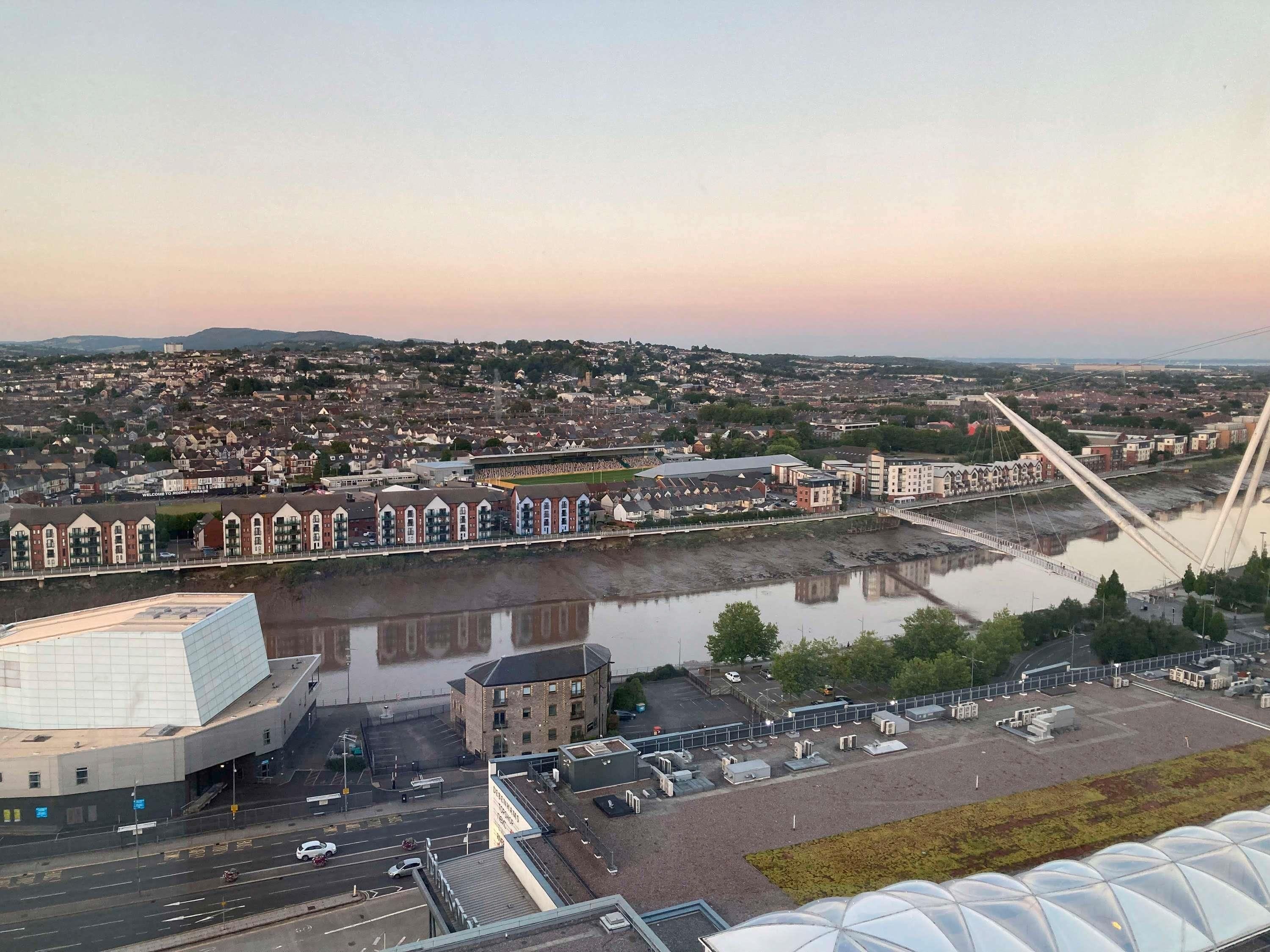 Ein modernes Gebäude mit einem einzigartigen geometrischen Dach, einem Parkplatz und Blick auf einen Fluss und eine Brücke.
