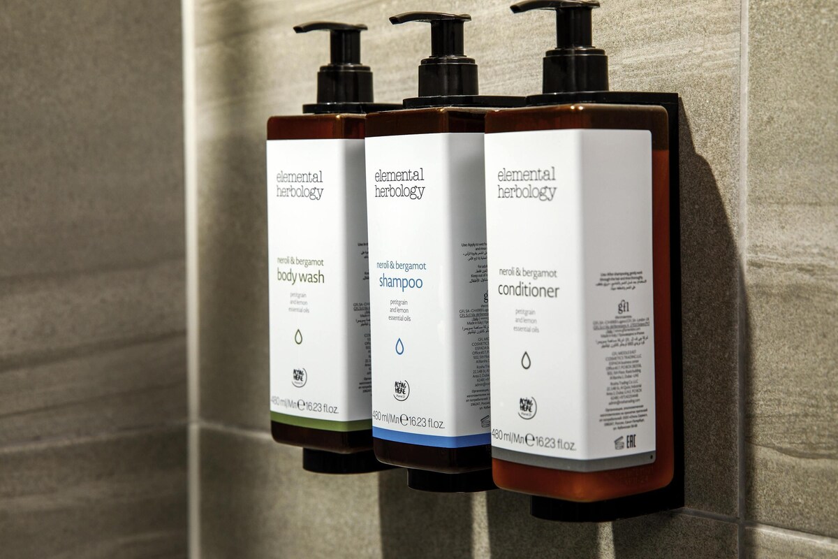 Three bottles of Elemental Herbology products: body wash, shampoo, and conditioner, each with a pump dispenser and labeled with volume and essential oils.