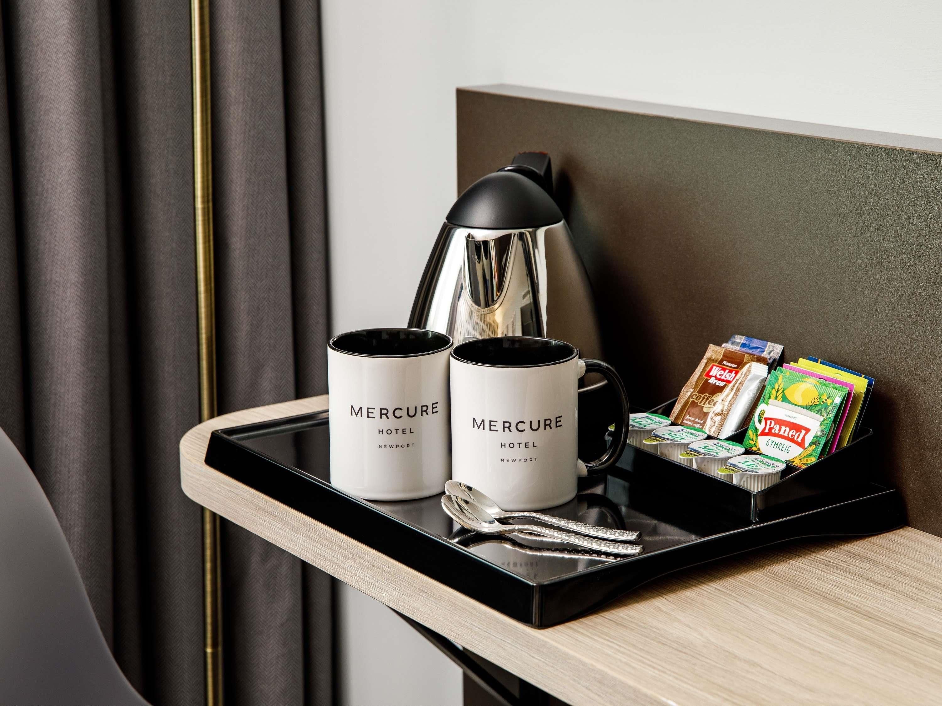 A tray with two mugs labeled 'Mercure Hotel Newport', a black lamp, and a tray of breakfast items including cereals and bread.