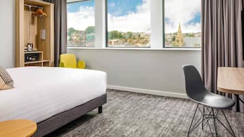 Habitación de hotel con cama, una silla, una mesa, armario y vista a la ciudad a través de grandes ventanales.