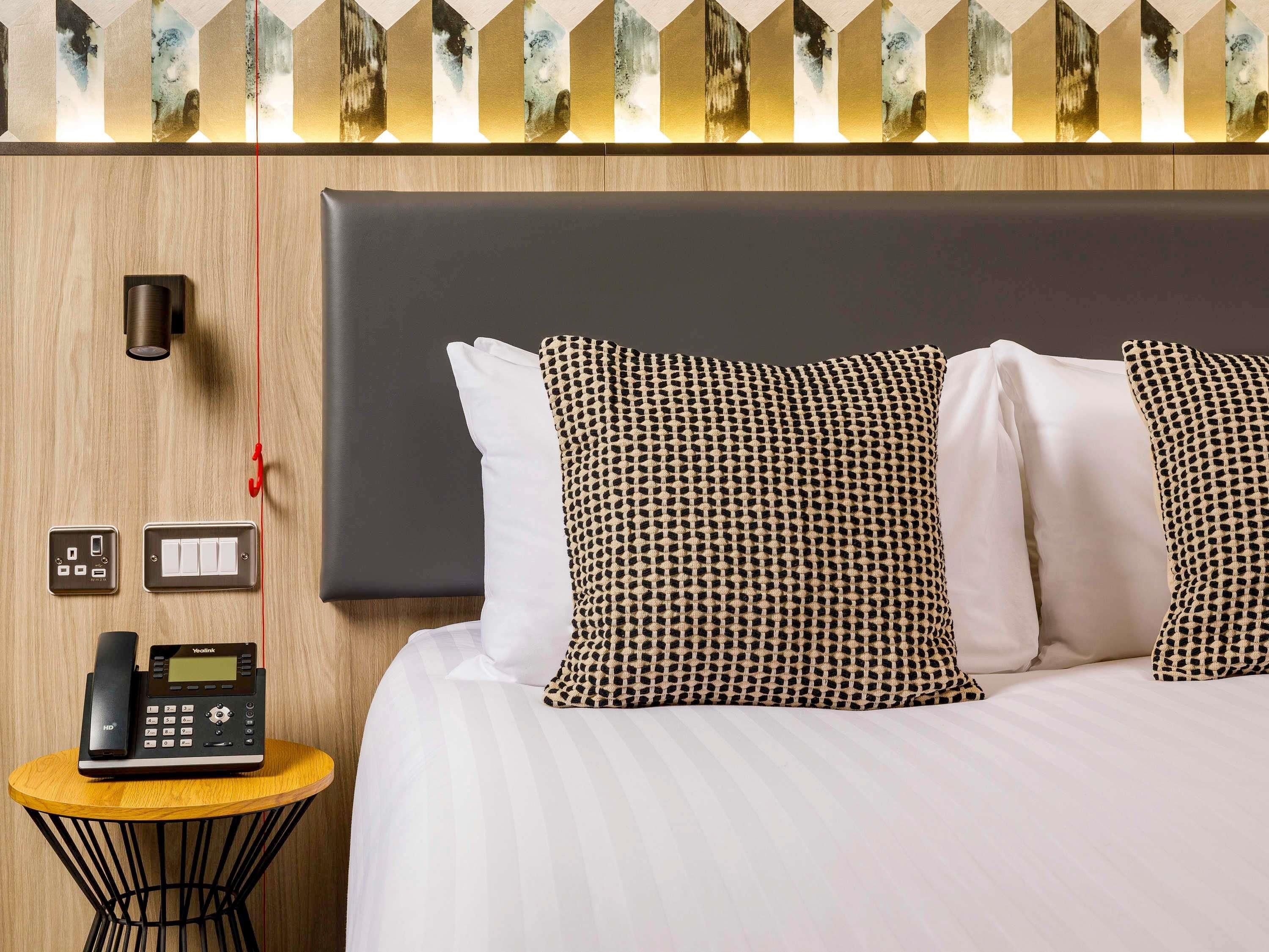 A hotel room with a bed, a bedside table with a phone, a wall-mounted light, and a power outlet.
